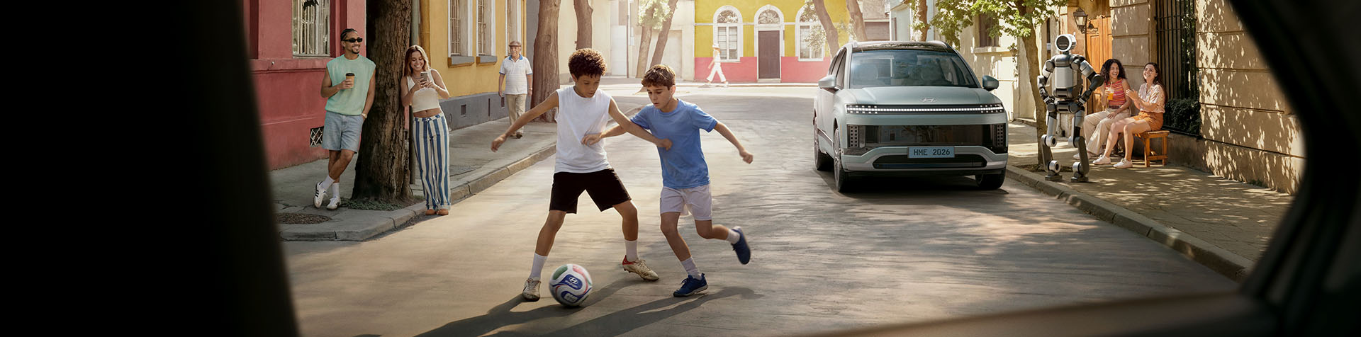 Two children playing football in a sunny residential street with a Hyundai IONIQ 9 and humanoid robot parked nearby while neighbours watch.