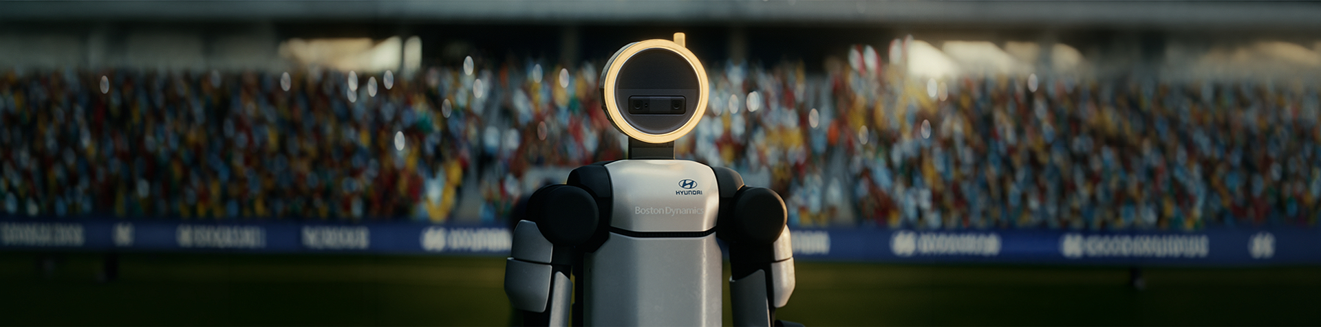 A humanoid robot stands on a stadium field, facing forward with a glowing circular sensor as its head, with a crowd of spectators in the background.