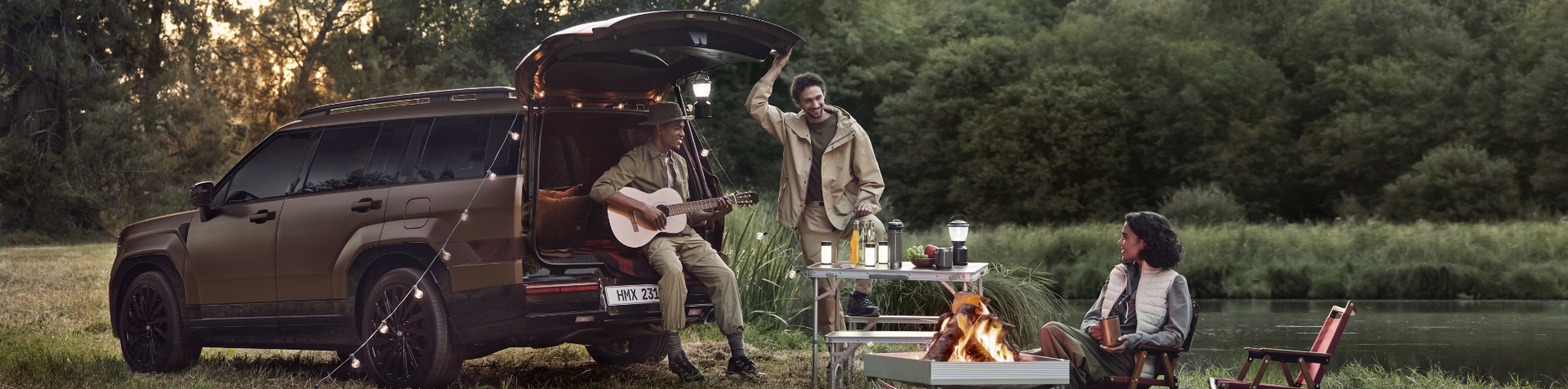 A parked Earthy Brass Metallic Matte colored The all-new SANTA FE with the tailgate open with a man sitting on the back playing guitar, a man standing to the side with his hand on the tailgate door, and a woman sitting by a campfire beside a camping table. 