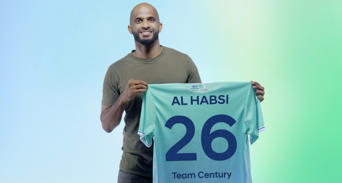 Torso view of new Team Century member Ali Al-Habsi smiling into the camera and holding up a green Team Century shirt with the number 26 and Team Century emblazoned in blue across the back.