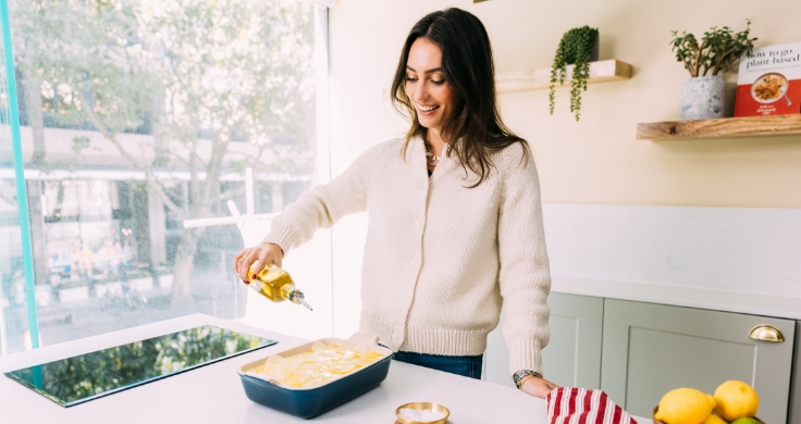 Ella Mills standing in her kitchen in front of a big window. She is drizzling olive oil on top of a dish that is ready to go into the oven. She is wearing a white cardigan and blue jeans.