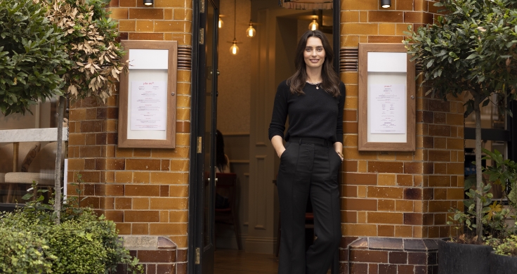 Ella Mills standing in the doorway of a restaurant. She has one hand on one side of the door frame, she has long brown hair and blue eyes and is wearing a black pullover and black pants.