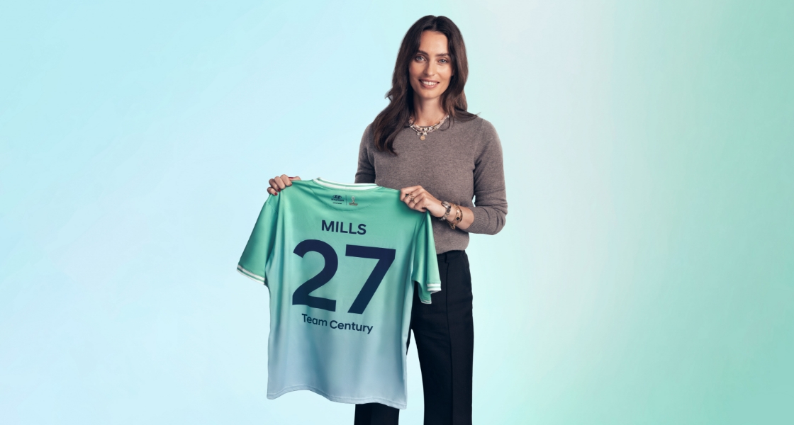 Ella Mills standing facing the camera with her Team Century jersey in her hands. She is wearing a gray pullover and black pants and her Team Century jersey is green and blue with Mills, 27 and Team Century written on it.